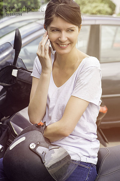 Frau sitzt auf einem geparkten Motorrad und redet mit dem Handy.