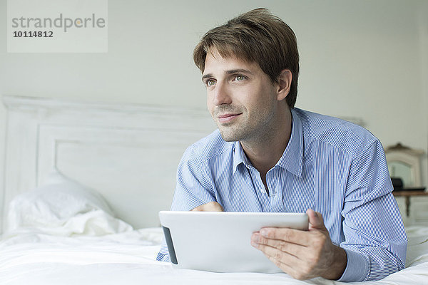 Mann im Bett liegend mit digitalem Tablett  wegblickend mit kontemplativem Blick