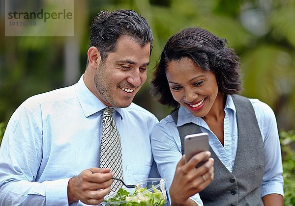 Geschäftsleute sitzen Seite an Seite und genießen einen Salat in der Mittagspause  schauen auf das Smartphone  lächeln