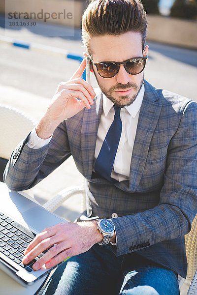 Stilvoller junger Geschäftsmann mit Smartphone und Laptop im Bürgersteigcafé