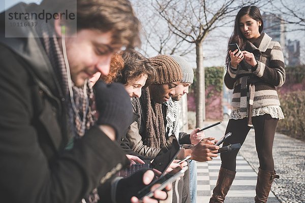 Sechs junge erwachsene Freunde mit Smartphones und digitalen Tabletts im Park