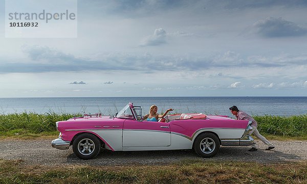 Junger Mann schiebt Vintage Cabrio an der Küste  während Freundin fährt  Havanna  Kuba