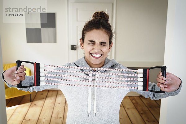 Junge Frau  die sich anstrengt  mit einem Brust-Expander im Wohnzimmer zu trainieren.