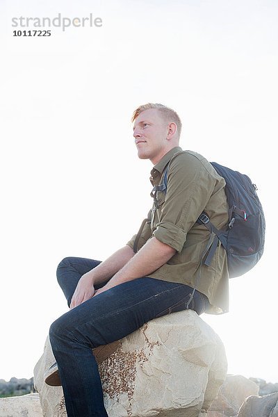 Junger Mann mit Rucksack auf einem Felsen sitzend  der wegguckt.