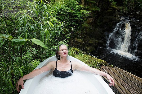 Reife Frau entspannt sich im Schaumbad vor dem Wasserfall beim Öko-Retreat