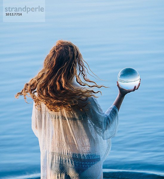 Rückansicht einer jungen Frau mit langen roten Haaren  die im See steht und eine Kristallkugel hält.