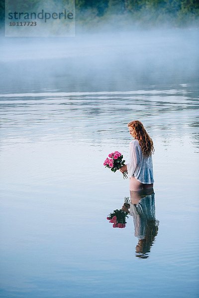 Junge Frau steht im nebligen See und hält Rosenstrauß