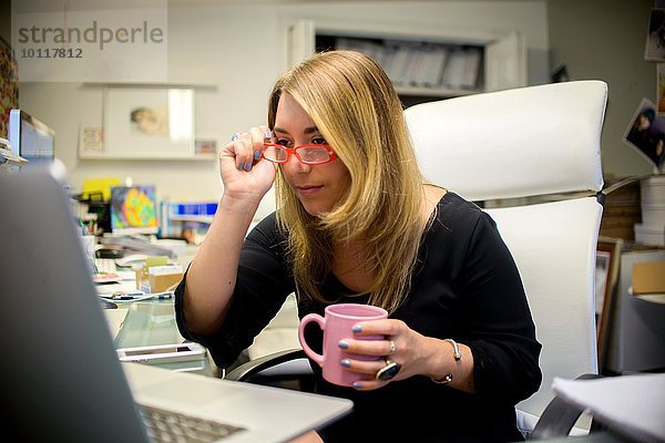 Junge Frau im Büro  am Schreibtisch sitzen  Kaffeetasse halten  Gläser aufsetzen