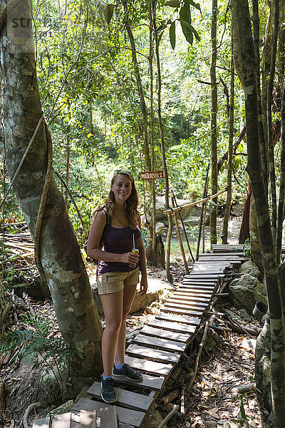 Touristin lächelt mit etwas zu trinken  auf einem Holzsteg im Wald  Weg zu den Na Muang Wasserfällen  Koh Samui  Thailand  Asien