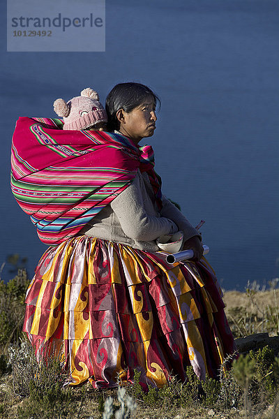Indiofrau mit Kleinkind auf dem Rücken in traditioneller Tracht vor dem Titikaka-See  Lago Titicaca  Sonneninsel  Isla del sol  Bolivien  Südamerika