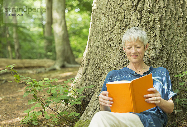 Lächelnde Seniorin liest Buch gegen Baum im Wald