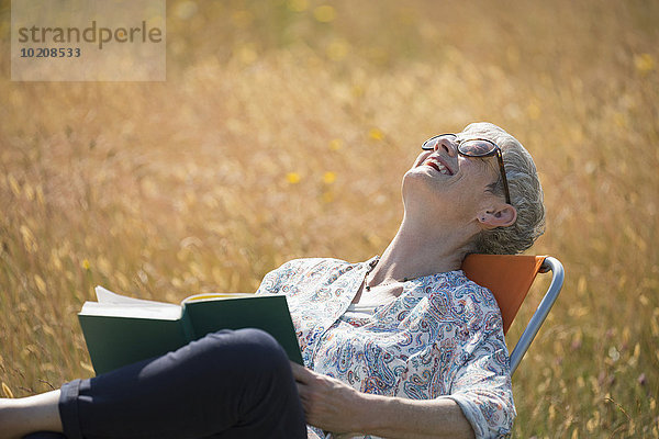 Seniorin liest Buch und lacht mit Kopf zurück im sonnigen Feld