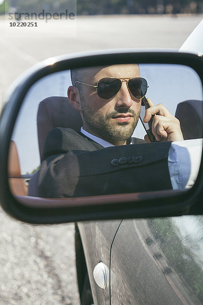 Spiegelbild des Geschäftsmannes mit Sonnenbrille beim Telefonieren im Auto