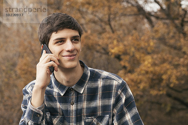 Porträt eines lächelnden jungen Mannes beim Telefonieren mit dem Smartphone im Park