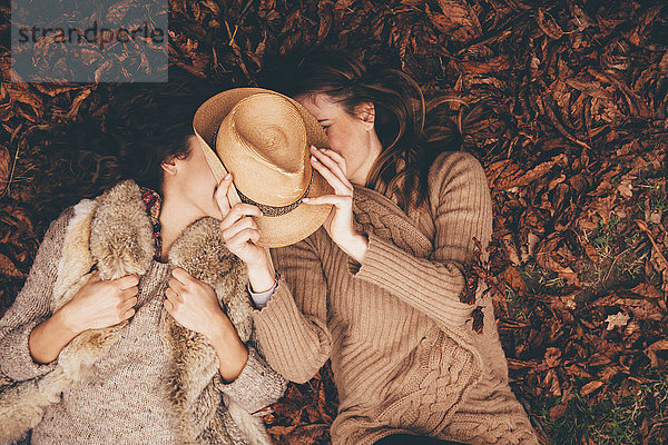 Zwei Freundinnen liegen Seite an Seite auf Herbstblättern in einem Park mit einem Hut.