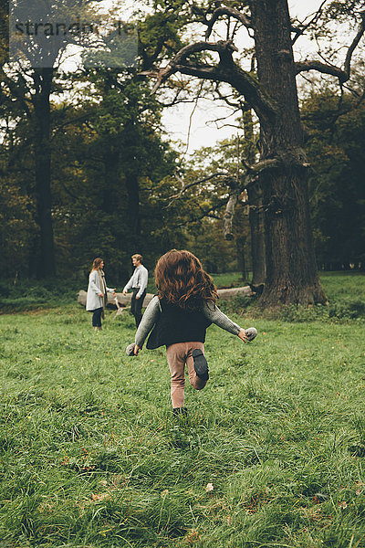 Rückansicht eines kleinen Mädchens  das in einem Park läuft  während seine Eltern im Hintergrund stehen.