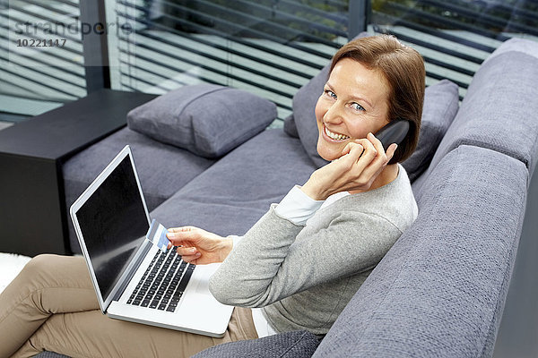 Porträt einer lächelnden Frau auf einer Couch mit Laptop und Kreditkarte beim Telefonieren