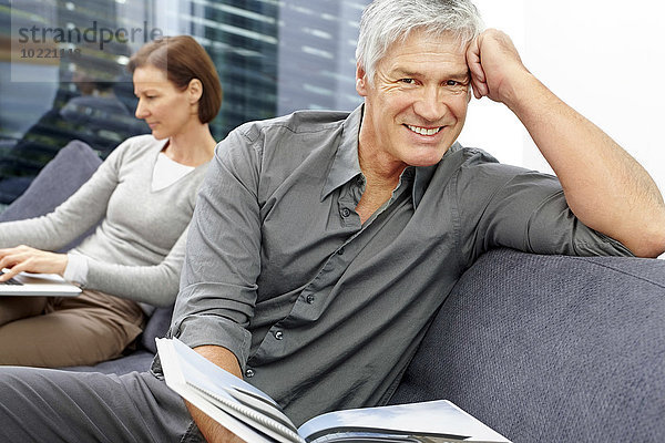Porträt eines entspannten Mannes  der mit einem Buch auf der Couch sitzt  während seine Frau im Hintergrund den Laptop benutzt.