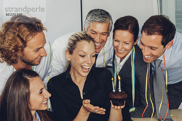Aufgeregte junge Geschäftsfrau hält einen kleinen Muffin mit brennenden Kerzen und feiert mit Kollegen ihren Geburtstag im Büro.