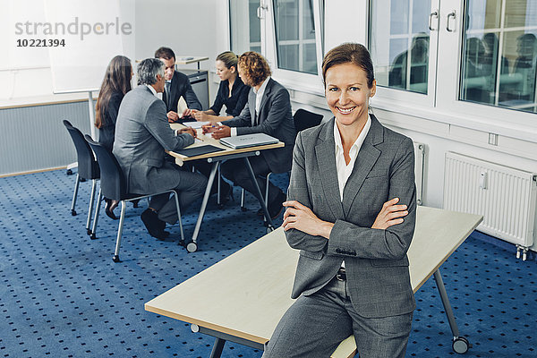 Reife Geschäftsfrau auf dem Schreibtisch sitzend mit gekreuzten Armen  Teamarbeit im Hintergrund