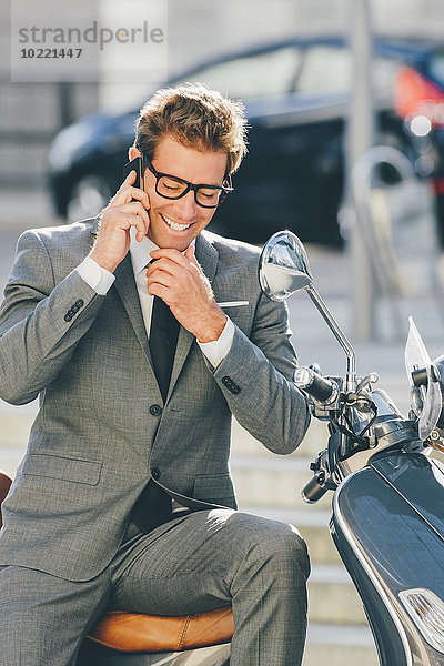 Lächelnder Geschäftsmann sitzt auf dem Motorroller und spricht auf dem Handy.