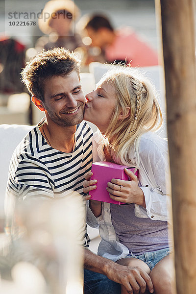 Junge Frau hält ein Geschenk und küsst ihren Freund im Outdoor-Café.