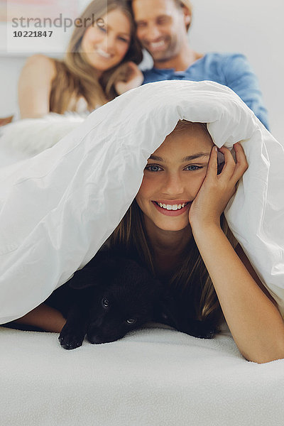Porträt eines glücklichen Mädchens mit Welpe im Bett und Eltern im Hintergrund