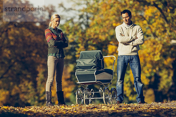 Reserviertes Paar steht mit gekreuzten Armen und schaut auf den Kinderwagen im Herbstpark.