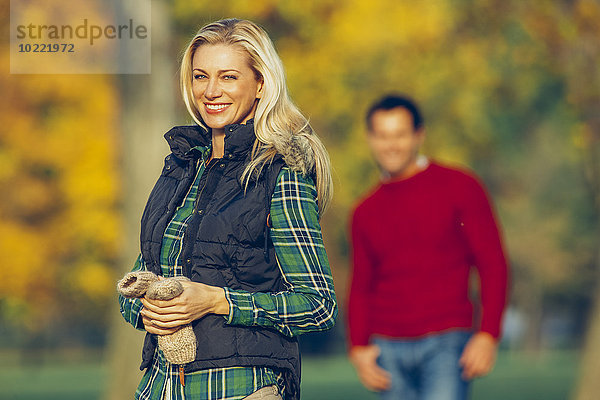 Porträt einer lächelnden  blonden Frau im herbstlichen Park mit einem Mann im Hintergrund.