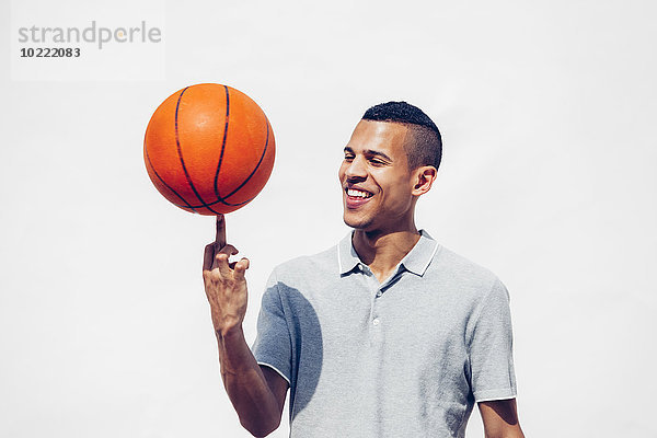 Porträt eines lächelnden jungen Mannes  der Basketball auf seinem Finger vor weißem Hintergrund balanciert.