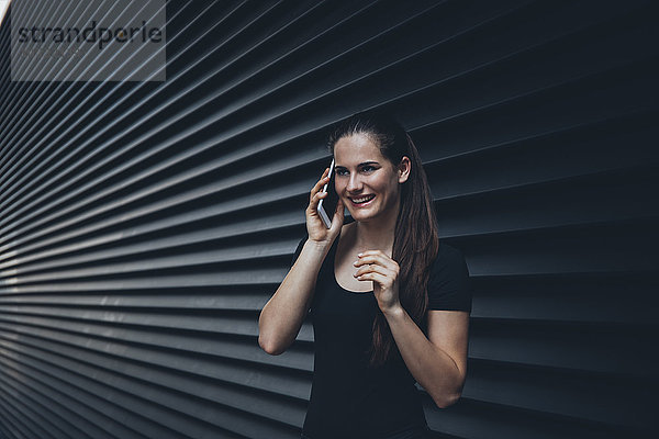 Porträt einer lächelnden jungen Frau beim Telefonieren mit Phablet vor schwarzer Fassade