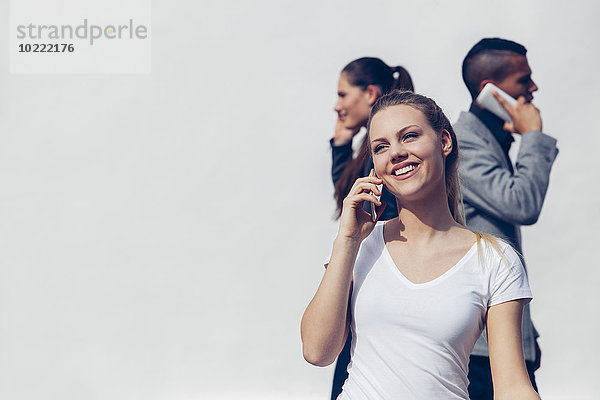 Porträt einer lächelnden jungen Frau  die mit dem Smartphone vor zwei anderen Menschen telefoniert.