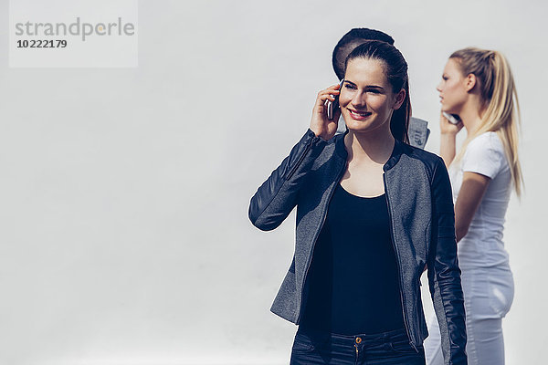 Porträt einer jungen Frau beim Telefonieren mit dem Smartphone vor zwei weiteren Personen