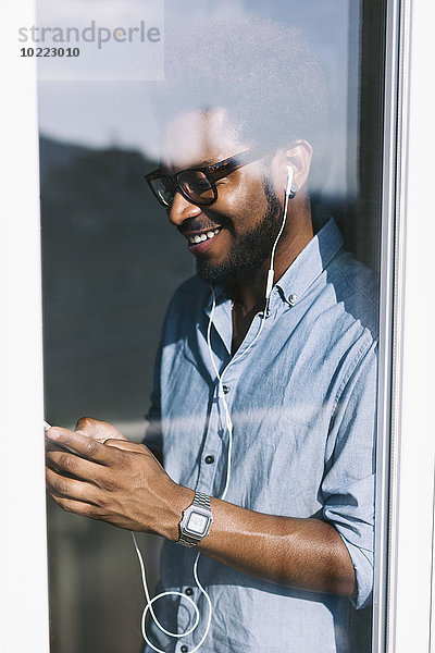 Lächelnder junger Mann hinter der Fensterscheibe hört Musik vom Smartphone