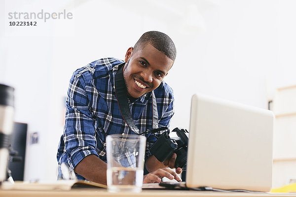 Porträt eines lächelnden jungen Mannes  der in seinem Fotostudio mit Laptop arbeitet.