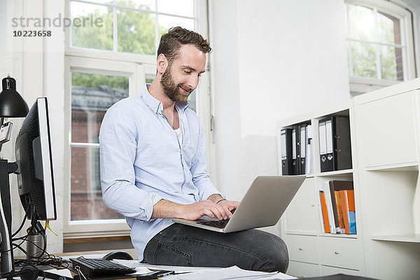 Lächelnder junger Mann im Büro sitzend auf dem Schreibtisch mit Laptop