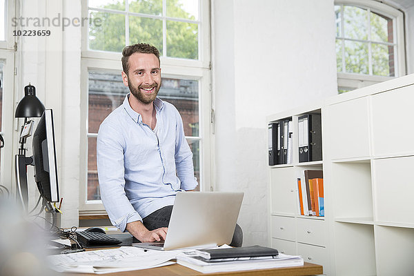 Lächelnder junger Mann im Büro am Schreibtisch mit Laptop