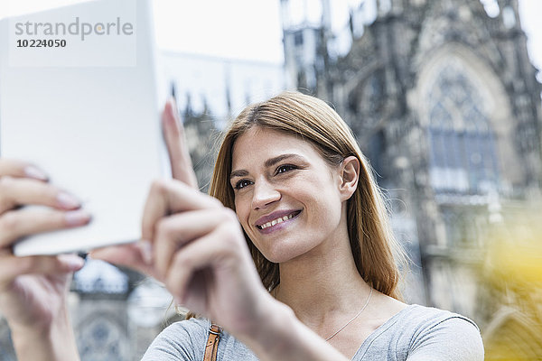 Deutschland  Köln  Porträt einer lächelnden jungen Frau  die vor dem Kölner Dom einen Selfie nimmt.