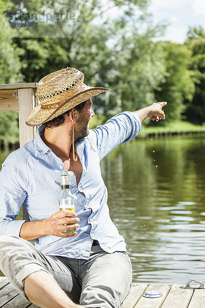 Mann sitzt auf der Plattform am Wasser mit einer Bierflasche  die mit dem Finger auf ihn zeigt.