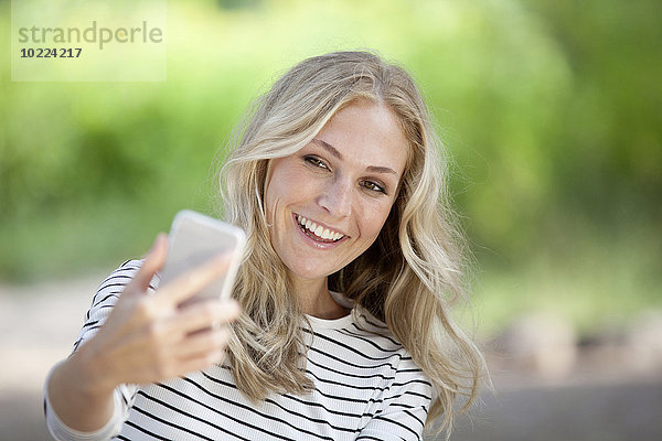Porträt einer glücklichen blonden Frau  die einen Selfie mit ihrem Smartphone nimmt.