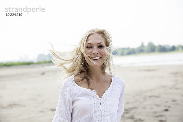 Porträt einer lächelnden blonden Frau mit blasendem Hait am Strand
