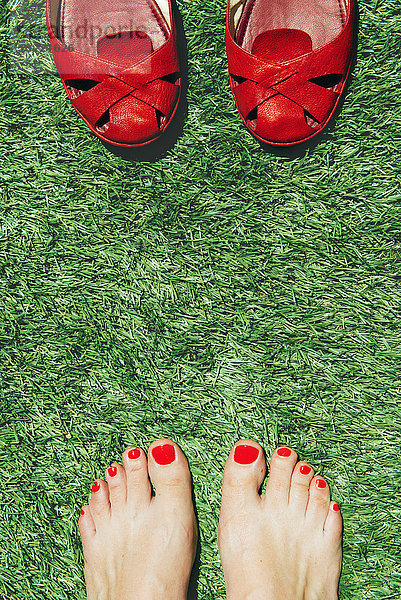 Barfuß-Frauen mit rot lackierten Nägeln neben einem Paar roter Schuhe  auf dem grünen Gras