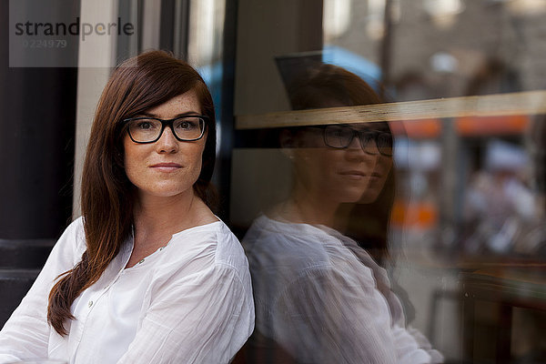 Porträt einer Frau mit braunen Haaren  die eine Brille trägt  die an der Fensterscheibe eines Cafés lehnt.