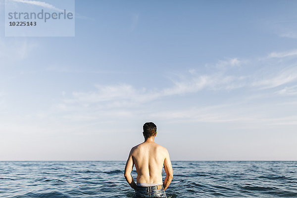 Rückansicht eines jungen Mannes  der im Meer steht und in die Ferne schaut.