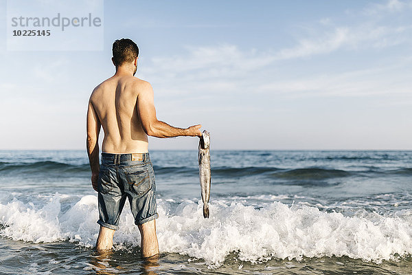 Rückansicht eines jungen Mannes  der im Wasser des Meeres steht und gefangene Fische hält.