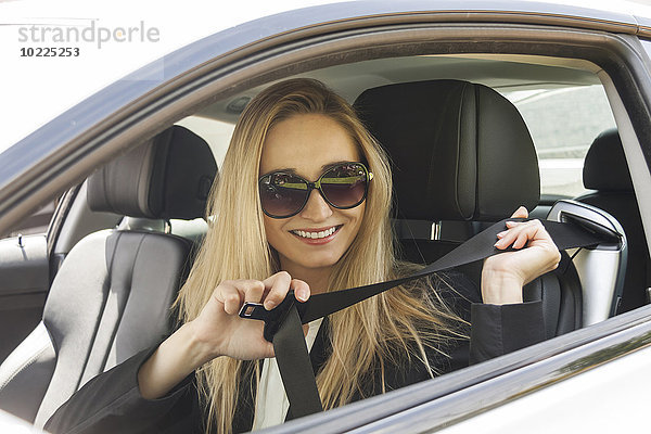 Porträt einer lächelnden blonden Frau mit Sonnenbrille  die sich im Auto anschnallt