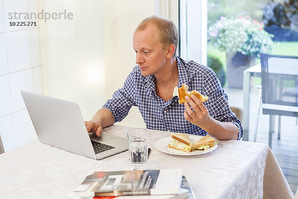 Mann zu Hause mit seinem Laptop beim Essen eines Snacks