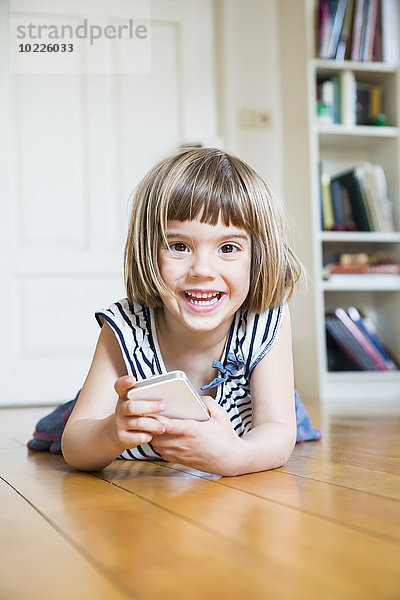 Portrait eines glücklichen kleinen Mädchens auf Holzboden liegend mit Smartphone
