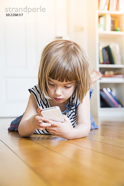 Kleines Mädchen auf Holzboden liegend mit Smartphone