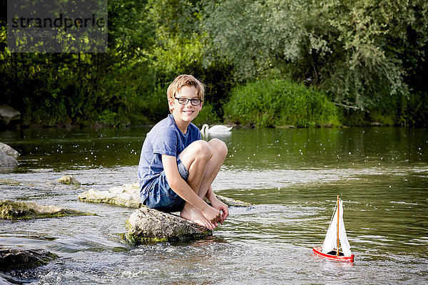 Junge spielt mit Holzspielzeugboot am Fluss